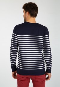 Langærmet skjorte i marineblå med hvide horisontale striber, lavet af en bomuldsblanding, med rund hals og tætsiddende design. Bæres med røde bukser.