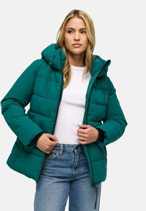 Femme aux longs cheveux blonds portant une doudoune bleu sarcelle par-dessus une chemise blanche et un jean bleu, tenant la fermeture éclair de la veste.