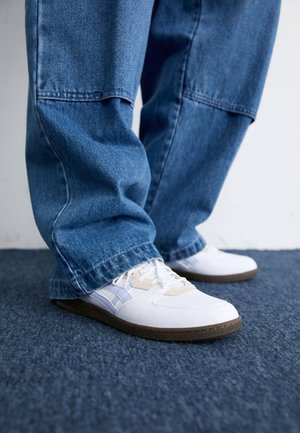 Person wearing loose blue jeans and white sneakers standing on blue carpeted floor.