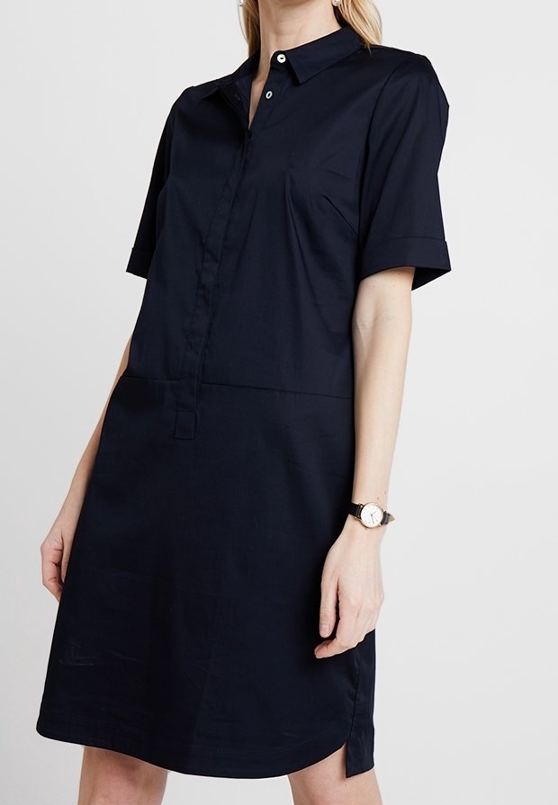 Donna che indossa un abito camicia blu navy a maniche corte e lunghezza al ginocchio, con colletto e un orologio al polso sinistro.