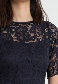 Marinblus med spets och blommönster, korta ärmar och rund halsringning. Har en texturerad, lager-på-lager design med genomskinliga detaljer.