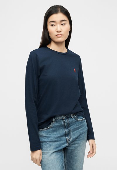 Νεαρή γυναίκα με μακριά σκούρα μαλλιά φοράει μπλε ναυτικό μπλουζάκι με μακριά μανίκια και μικρό κόκκινο λογότυπο, καθώς και ανοιχτόχρωμο μπλε τζιν, κοιτάζοντας προς τα πλάγια.