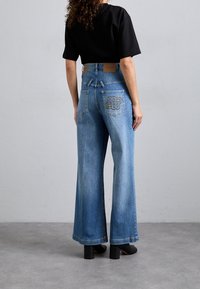 Femme portant un jean bleu taille haute à jambes larges avec une poche arrière brodée et un haut noir à manches courtes, debout sur un sol gris.