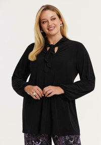 Smilende kvinde med langt blond hår har en sort bluse med sløjfe ved halsen og pufærmer, parret med mørke bukser med blomsterprint.