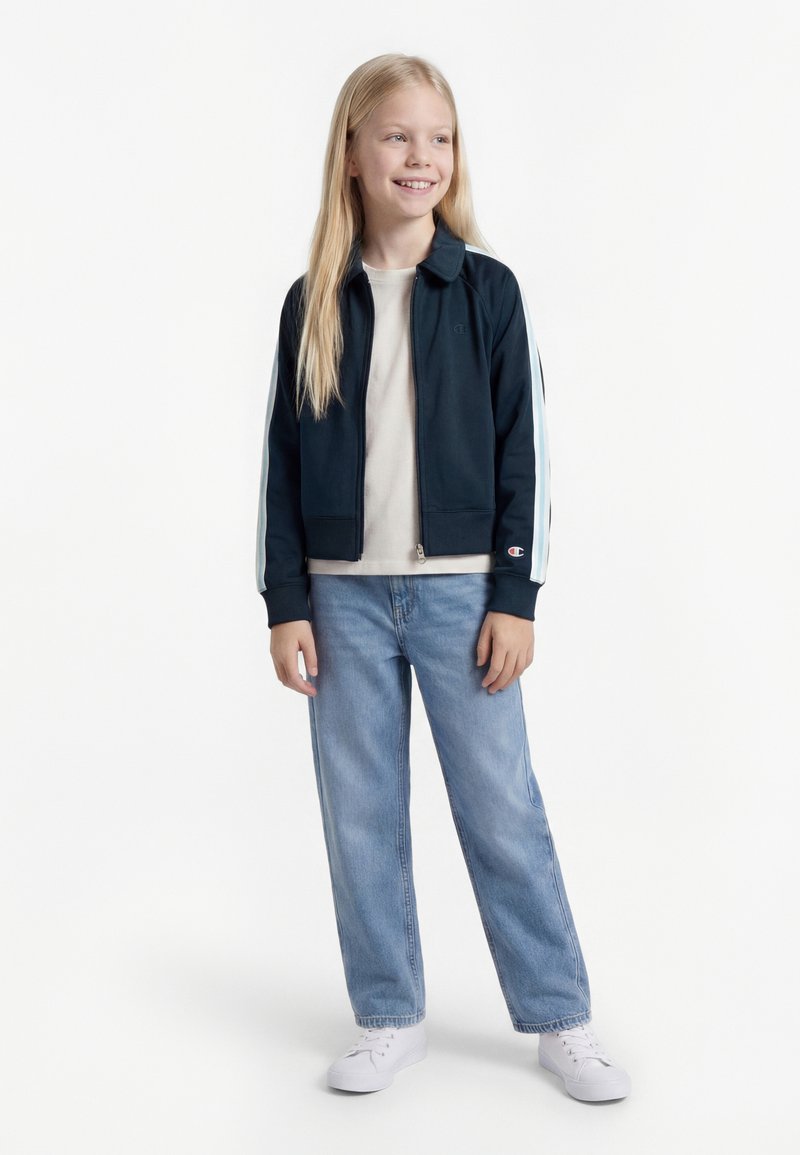 Mladé dievča sa usmieva, má na sebe námornícky modrú bundu s bielymi pruhmi, béžové tričko, svetlomodré džínsy a biele tenisky, stojí na jednoliatom pozadí.