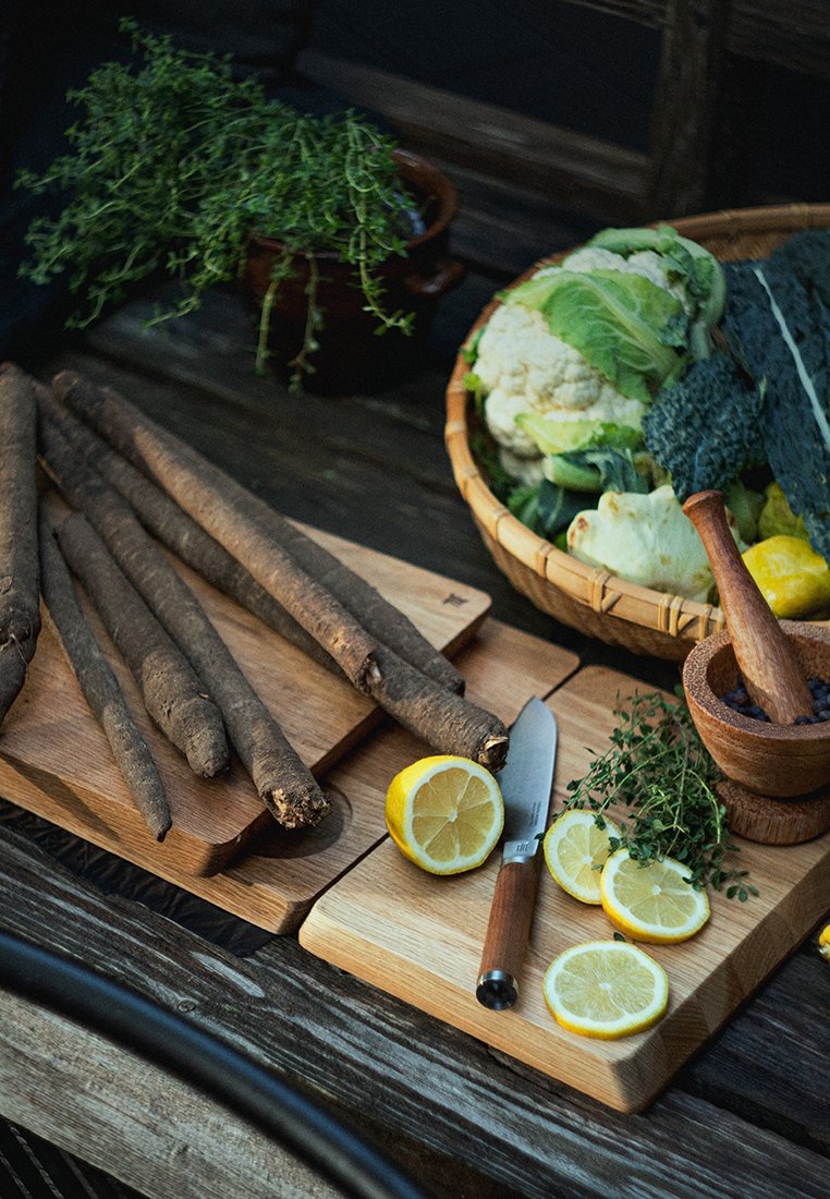 Korzenie salsify na drewnianych deskach, pokrojone cytryny, zioła, rustykalny nóż i kosz z mieszanymi warzywami ułożone na ciemnej drewnianej powierzchni.