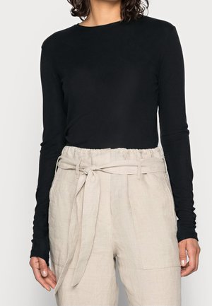 Kobieta ubrana w dopasowaną czarną bluzkę z długim rękawem, włożoną do wysokich beżowych spodni typu paperbag z wiązanym materiałowym paskiem.