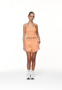 Oranžový rebrovaný crop top s tenkými ramienkami a bielym logom, spárovaný s nohavicami na zodpovedajúcom základe, ktoré majú elastický pás a vrecká.