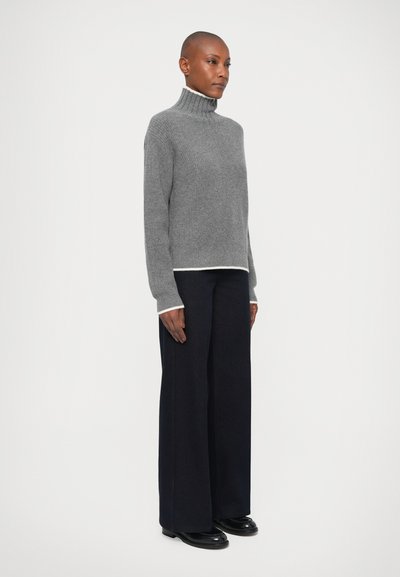 Pull en col roulé gris avec une finition texturée et un ourlet blanc contrastant, associé à un pantalon large bleu marine et des chaussures noires.