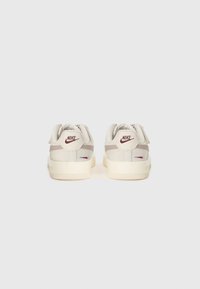 Baskets Nike avec une tige crème et des accents gris clair, une semelle plate, un logo à l'arrière et deux straps réglables pour le maintien.
