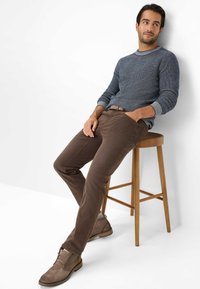 Chinos marroni scuri con una finitura testurizzata, abbinati a un maglione in maglia blu navy con colletto grigio chiaro. Stivaletti alla caviglia in suede marroni.