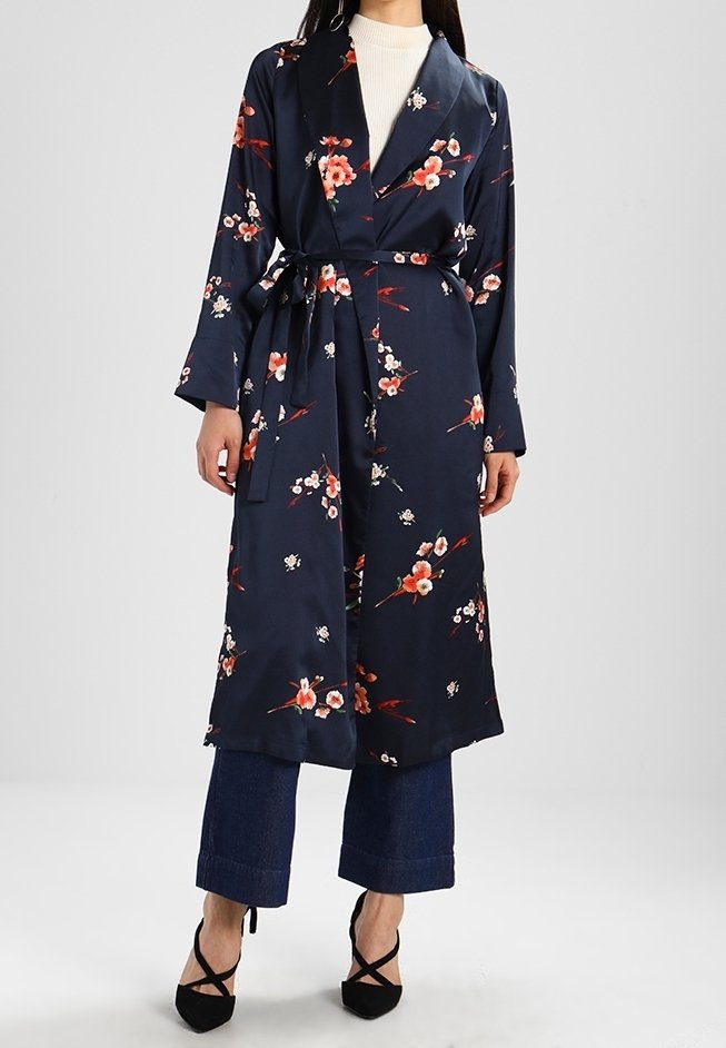 Femme portant un manteau portefeuille bleu marine à fleurs sur un haut blanc et un pantalon court bleu foncé avec des talons noirs pointus.