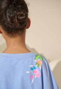 Világoskék pamut póló színes virágmintával a bal vállnál, kerek nyakkivágással és rövid ujjakkal. Puha tapintású anyag.