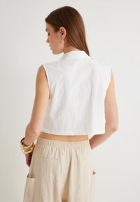 Top corto bianco senza maniche con un design texturizzato, dotato di un colletto classico, abbinato a pantaloni elasticizzati beige chiaro in vita. Bracciali d'oro al polso.