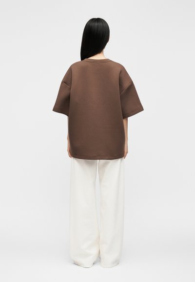 Γυναίκα με μακριά μαύρα μαλλιά φοράει υπερμεγέθη καφέ κοντομάνικη μπλούζα και φαρδιές λευκές παντελόνες, στέκεται με την πλάτη στον απλό φόντο.