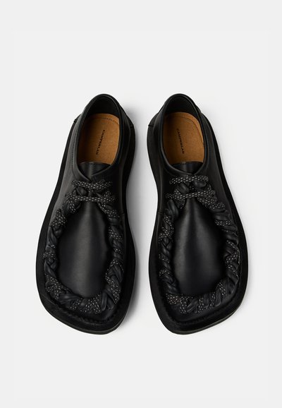 Chaussures en cuir noir à enfiler avec un détail en corde tressée le long du bord avant et des semelles intérieures rembourrées couleur beige sur fond blanc.