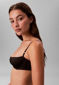 Czarny biustonosz typu balconette z cienkimi regulowanymi ramiączkami, gładką satynową teksturą, usztywnianymi miseczkami oraz detalami na szwach bocznych, na neutralnym tle.