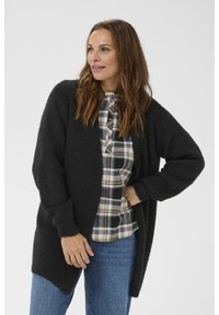 Zwarte gebreide cardigan met een relaxte fit, met lange mouwen en een gestructureerd oppervlak, gedragen over een geruite overhemd met knopen.