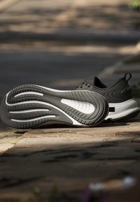 Chaussure de sport noire et blanche posée sur un sol pavé, semelle visible avec des motifs incurvés et un design texturé sur la partie supérieure.