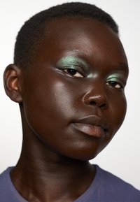 Gros plan sur un visage avec une peau lisse et foncée, portant un fard à paupières vert et une couleur de lèvres neutre, avec des cheveux courts et texturés, et un t-shirt violet.