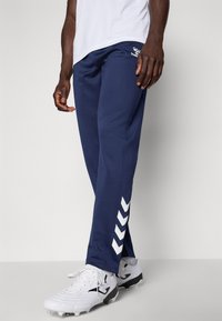 Muškarac u tamnoplavim sportskim hlačama s bijelim cik-cak prugama na donjem dijelu noge i bijelim kopačkama stoji ispred jednostavne pozadine.