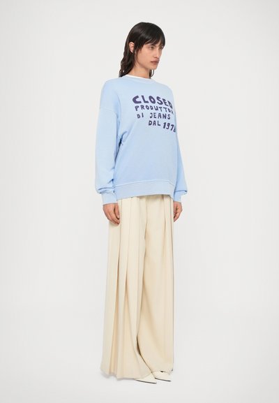 Ανοιχτό μπλε φούτερ με έντονη σκούρα στάμπα, χαλαρή εφαρμογή, ριμπ μανσέτες. Συνδυασμένο με εκρού παντελόνι φαρδιάς γραμμής και λευκά μυτερά παπούτσια.