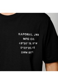 T-shirt en coton noir avec texte imprimé blanc : "KAPORAL JNS MFG CO.", coordonnées, et "DNM DEPT". Manches courtes, encolure ronde.