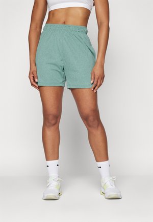 Kobieta w zielonych sportowych szortach, białym sportowym biustonoszu, białych skarpetkach Nike oraz białych butach sportowych stoi na jednolitym tle.