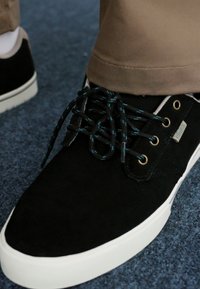 Crne suede tenisice s bijelim gumenim potplatom, s tamno tirkiznim vezicama, mesinganim rupicama i minimalističkim logotipom na strani.