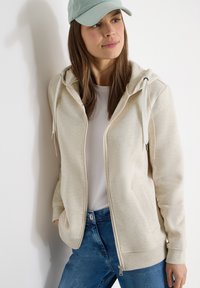 Femme portant un sweat à capuche zippé beige clair, un t-shirt blanc, un jean bleu et une casquette vert clair, debout contre un mur blanc.