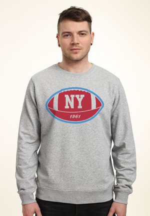 Mężczyzna w szarej bluzie z czerwono-niebieską grafiką piłkarską i napisem "NY 1961", stojący na jasnym tle.