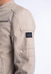 Beige jack met zachte textuur, voorzien van een ritszak, schoudernaden en een zwart merkpatch op de mouw.