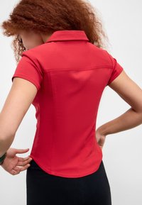 Polo shirt ajusté rouge à manches courtes et col. Texture lisse et silhouette cintrée. Présente un ourlet arrondi et des détails sur les côtés.