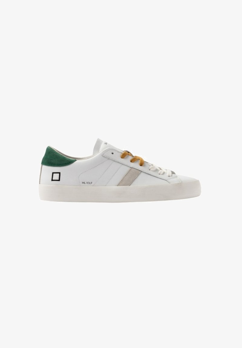 Witte lage sneaker met beige streep, groene hieltab, mosterdbotten en witte veters, en zwart vierkant logo aan de zijkant.