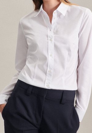 Kobieta ubrana w dopasowaną białą koszulę na guziki wpuszczoną do wysokich czarnych spodni, stoi z rękami w kieszeniach.