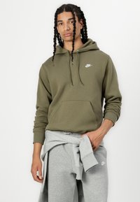 Κυπαρισσί πράσινο φούτερ Nike με μπροστινή τσέπη, κουκούλα με κορδόνι και μικρό λευκό λογότυπο. Συνδυάζεται με γκρι αθλητική φόρμα, επίσης με λογότυπο.