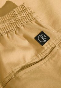 Tejido beige con una textura suave y una cinturilla elástica. Presenta una etiqueta cuadrada negra con un logo circular cosido en el bolsillo.