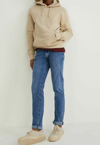 Un hoodie beige avec poche et cordons, superposé à une chemise bordeaux. Un jean en denim bleu avec les poignets retroussés, associé à des baskets beiges.