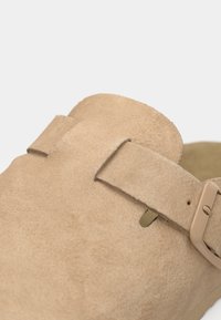 Beige velúr szandál széles pánttal és állítható csattal. Texturált felszín kerek orral és lapos talppal. Egyszerű dizájn, minta nélkül.