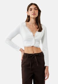 Biały, prążkowany, długi rękaw crop top z koronkowym wykończeniem, z guzikiem z przodu i dekoltem w serek. W zestawie z brązowymi spodniami z troczkami.