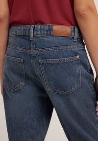 Jeans de mezclilla azul oscuro con una etiqueta de cuero en la cintura y dos bolsillos traseros, con detalles de costura y una textura ligeramente desgastada.