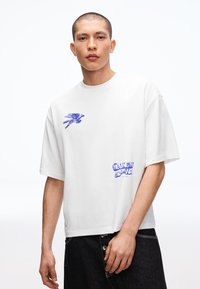 Fiatal férfi, aki fehér, túlméretezett pólót visel kék madár grafikával és "MOVING MY OWN WAY" felirattal, fehér háttér előtt áll.