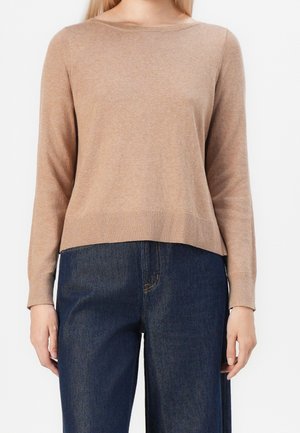 Vrouw draagt een beige trui met lange mouwen en geribbelde zoom, gecombineerd met donkerblauwe high-waisted spijkerbroek, staand tegen een witte achtergrond.