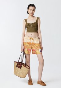 Top cortissimo verde oliva con bottoni, pantaloni multicolore con tie-dye, borsa a tracolla di paglia con dettagli in pelle scura e scarpe marroni slip-on.