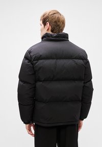 Crna puf jakna s visokim ovratnikom, s kvadratnim sekcijama i sužavajućim donjim rubom. Izrađena od glatkog, vodootpornog materijala.