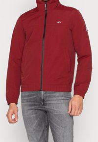 Rote, leichte Jacke mit hohem Kragen, Reißverschluss, zwei Seitentaschen und elastischen Bündchen. Mit kleinem Logo auf der Brust.