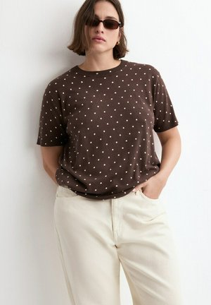 Femme portant une chemise marron à manches courtes à pois, un pantalon crème, des lunettes de soleil foncées et des boucles d'oreilles créoles en or, debout les mains dans les poches.