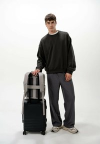 Valigia nera con ruote, dalla superficie texture e con spallacci grigi. Il modello indossa un maglione scuro e pantaloni grigi, in piedi su uno sfondo chiaro.