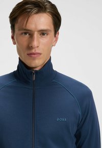 Giovane uomo con pelle chiara e capelli castani che indossa una giacca BOSS blu navy con zip e colletto alto, rivolto in avanti su uno sfondo semplice.