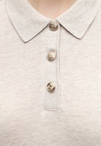 Polo beige avec trois boutons métalliques gravés d'un blason, gros plan sur le col et la patte de boutonnage.